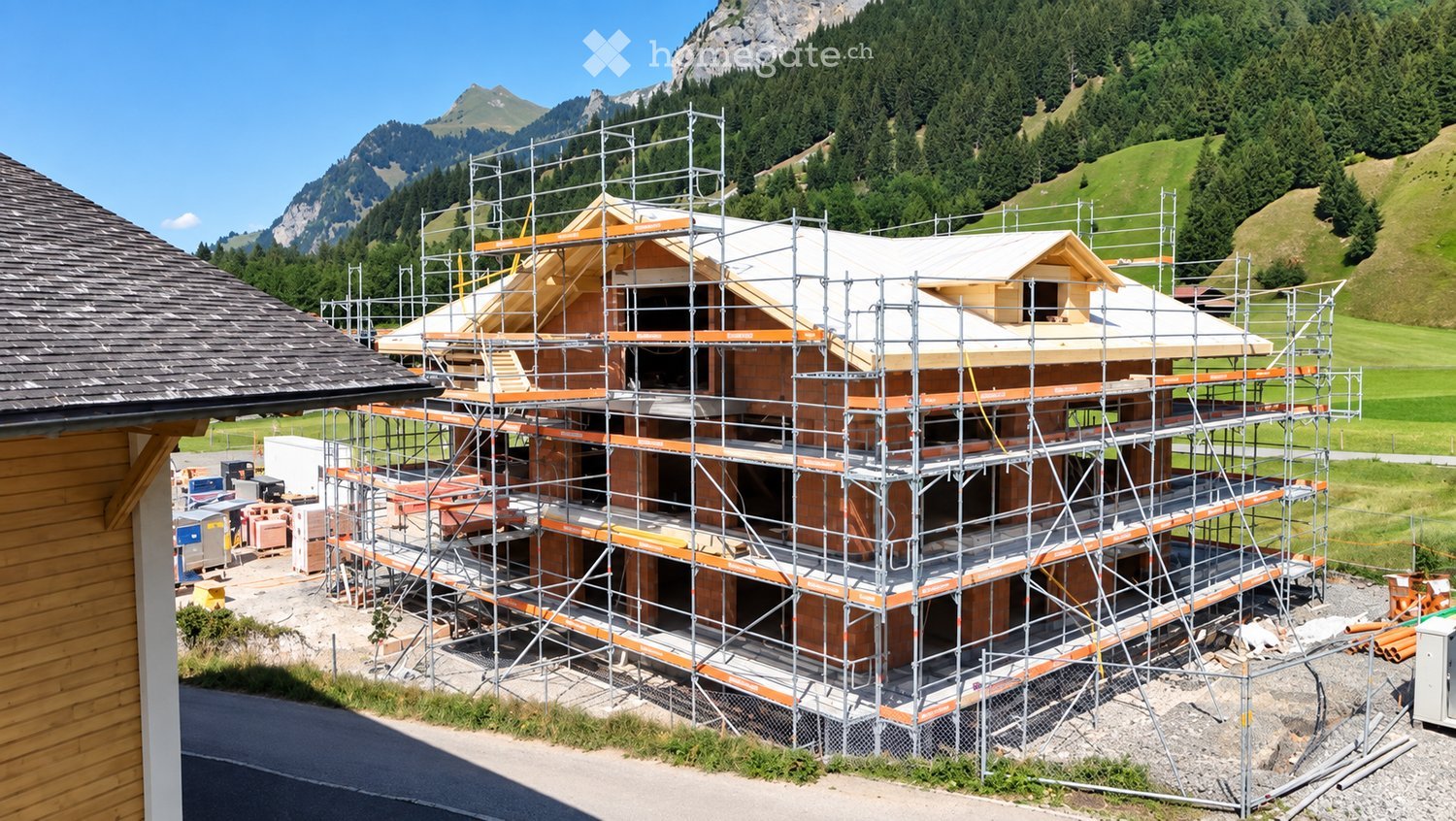 A multi-story house under construction, covered in scaffolding, with visible wooden beams and brickwork. The surrounding area features a grassy field and mountain backdrop.