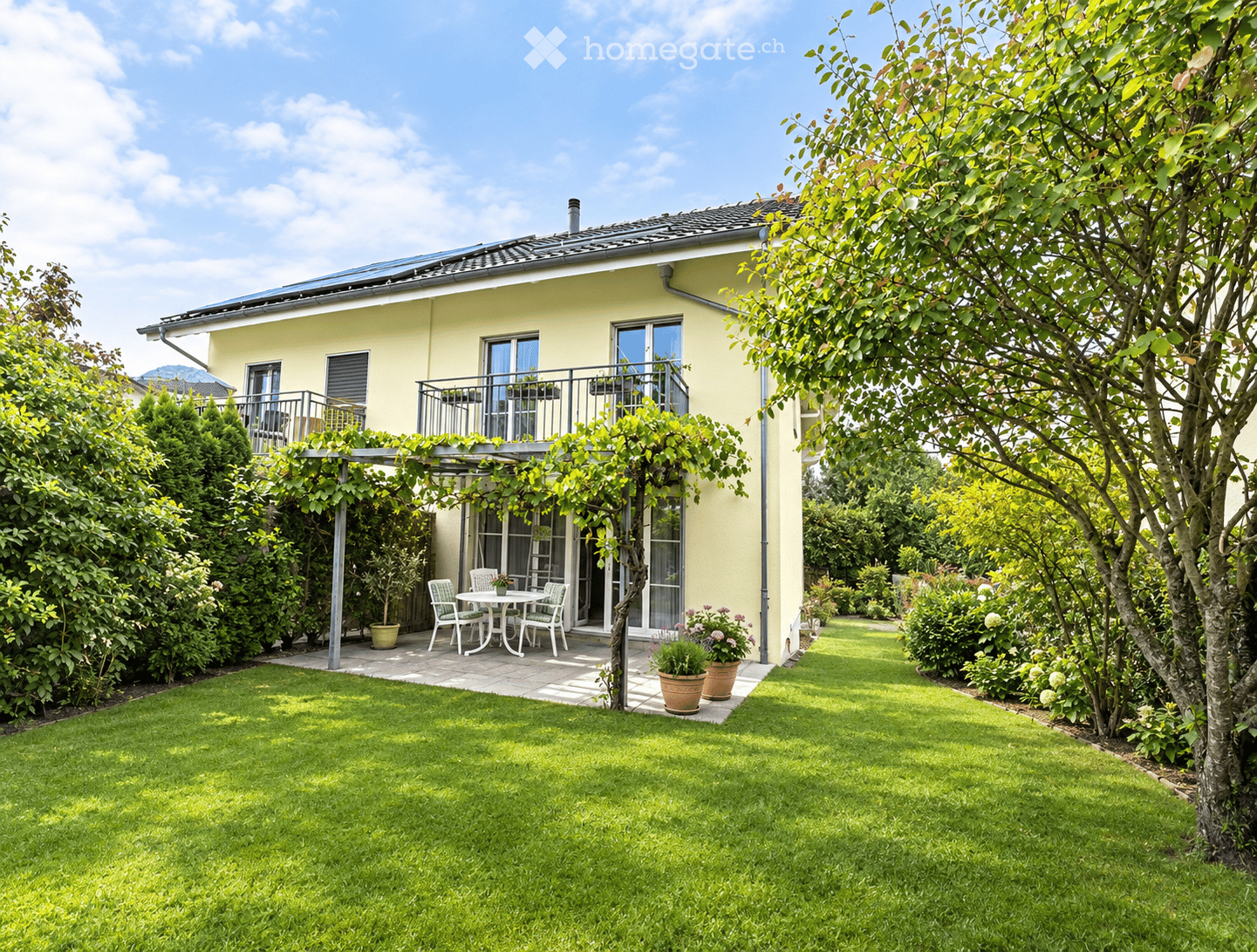A well-maintained garden with a lawn, a patio area with a table and chairs, potted plants, and a house with a balcony and large windows.