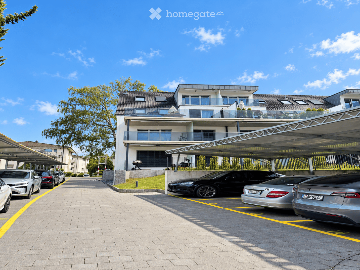 Modern multi-story residential building with balconies, surrounded by a paved parking area with several parked cars under covered structures.