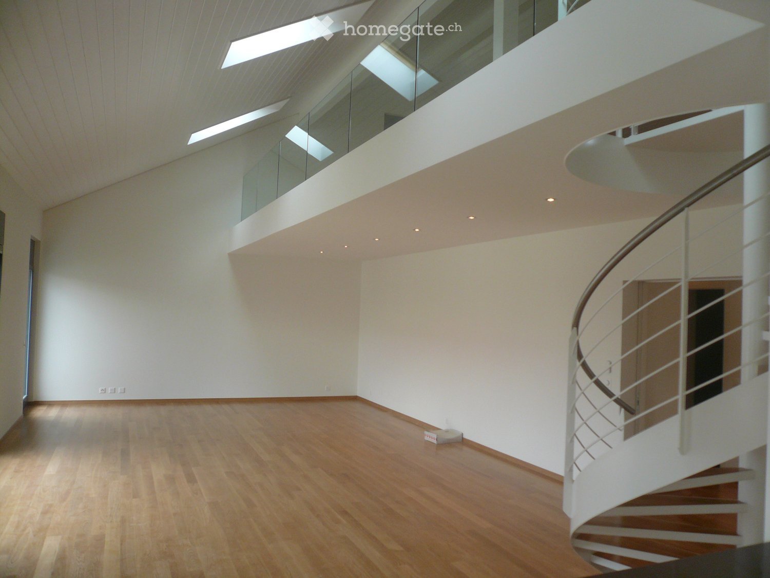 Empty room with wooden flooring, white walls, and a modern spiral staircase with a glass railing. The room features recessed lighting and a large skylight above.