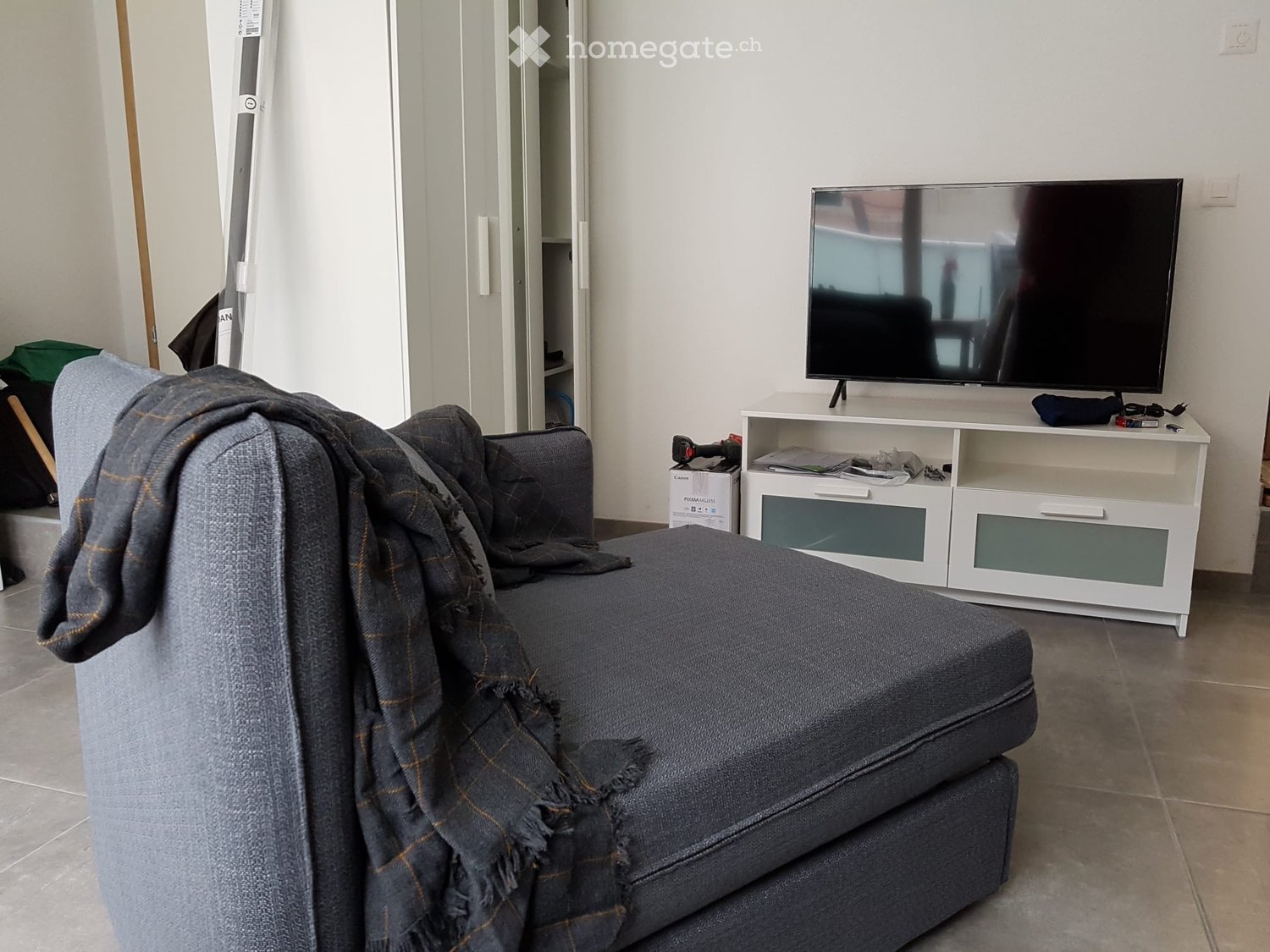 Living room with a grey sofa draped with a blanket, a flat-screen TV on a white cabinet, and a closet in the background.