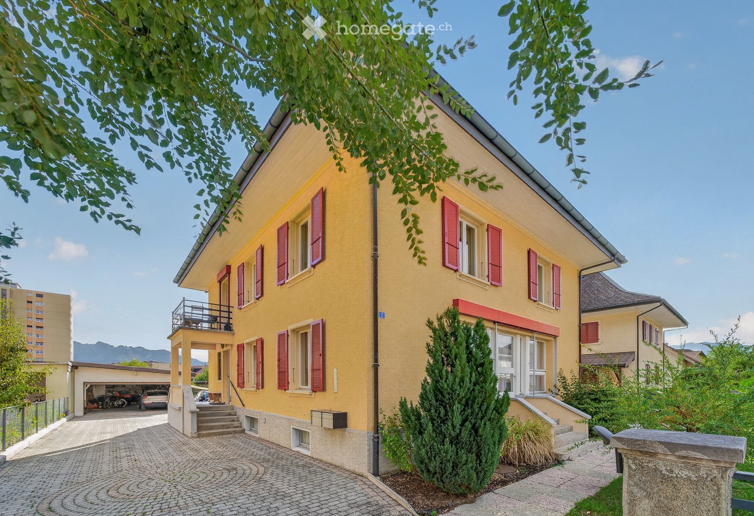 Two-story yellow house with red shutters, a balcony, and a garage. The house is surrounded by greenery and has a cobblestone driveway.