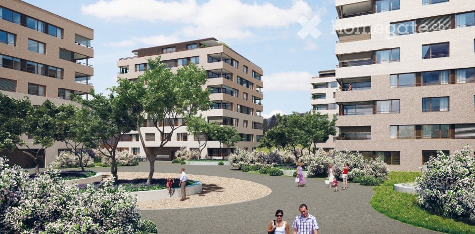 A modern residential complex with multiple apartment buildings, surrounded by landscaped gardens and pathways. The buildings feature contemporary architecture with large windows and balconies. The area includes trees, shrubs, and a central paved walkway w
