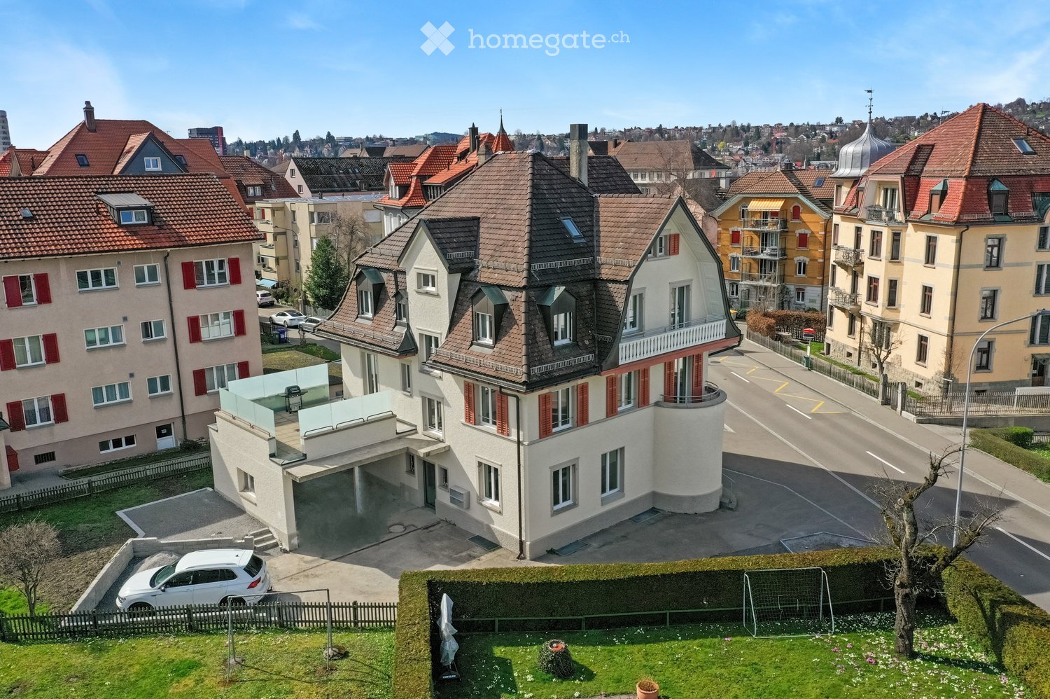 A multi-story house with a red-tiled roof, white walls, and red shutters. The house features a modern garage and a balcony on the upper floor. The surrounding area includes other residential buildings, a driveway, and a small garden with a tree and a socc