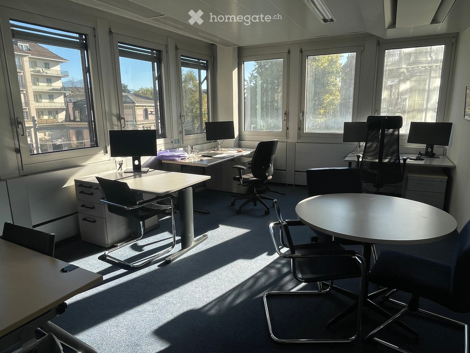 Bright office space with large windows, desks, chairs, computer monitors, and a round meeting table.