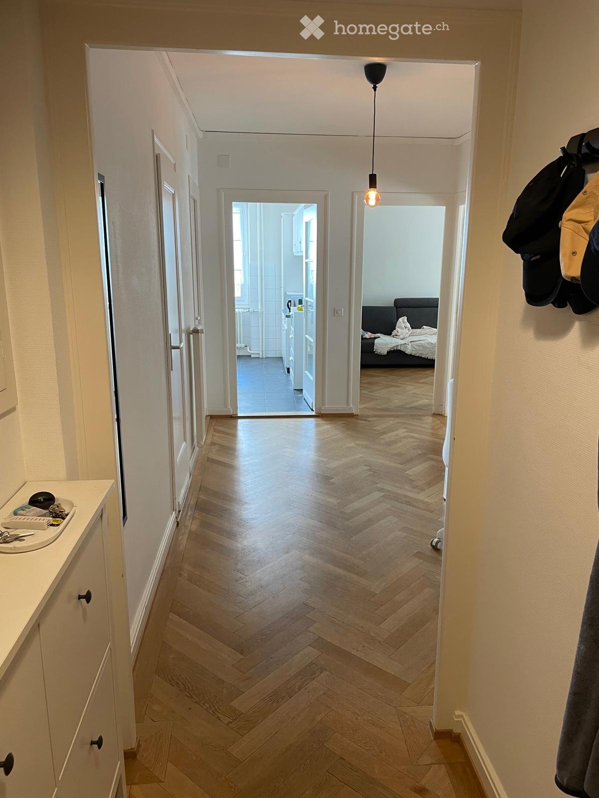 A well-lit hallway with herringbone wooden flooring, leading to a bedroom and a kitchen area. Visible items include a coat rack with jackets, a small sink with toiletries, and a hanging light fixture.