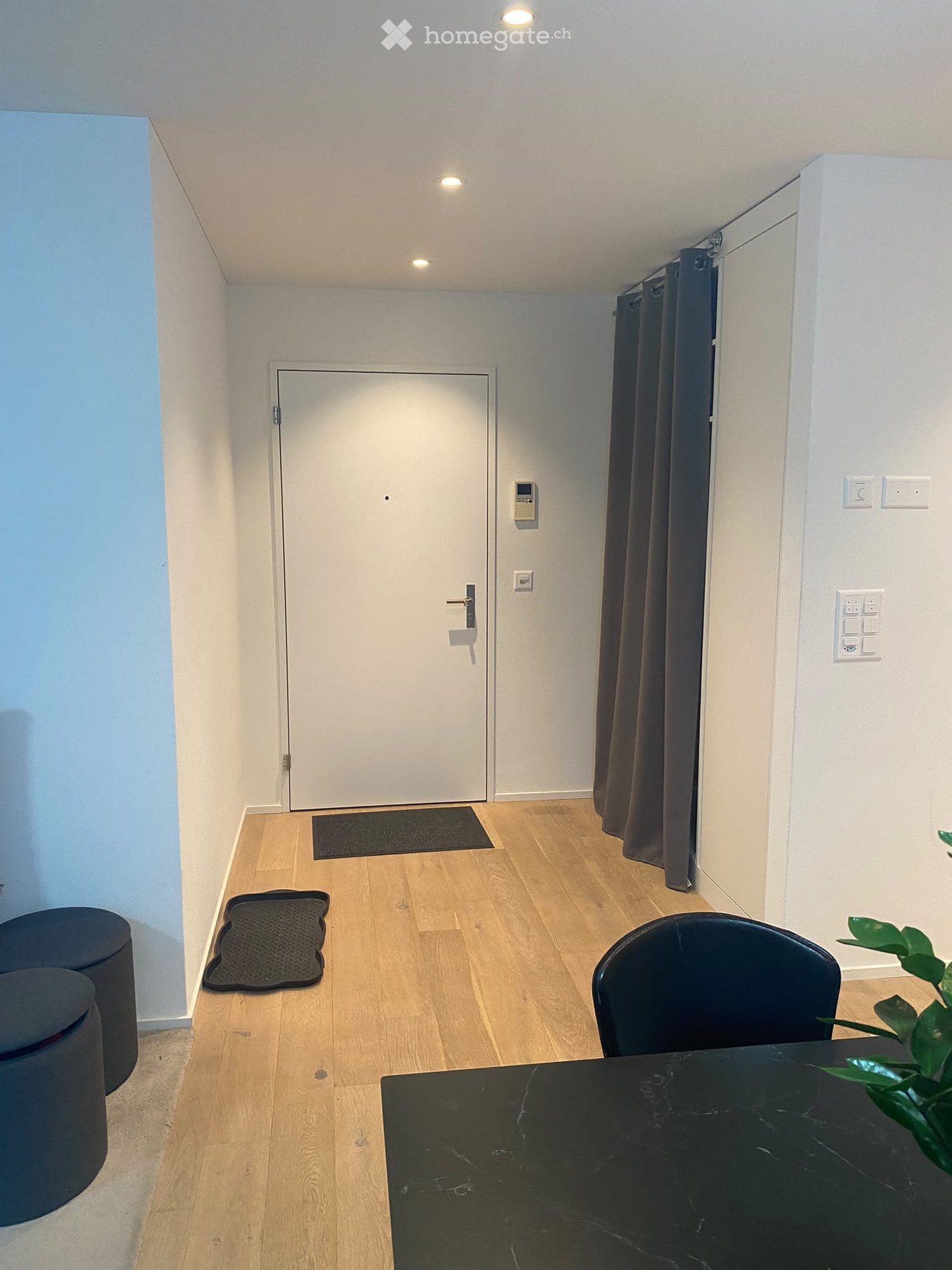 A small, modern hallway with wooden flooring, a black table, a chair, two stools, a door, and a curtain.