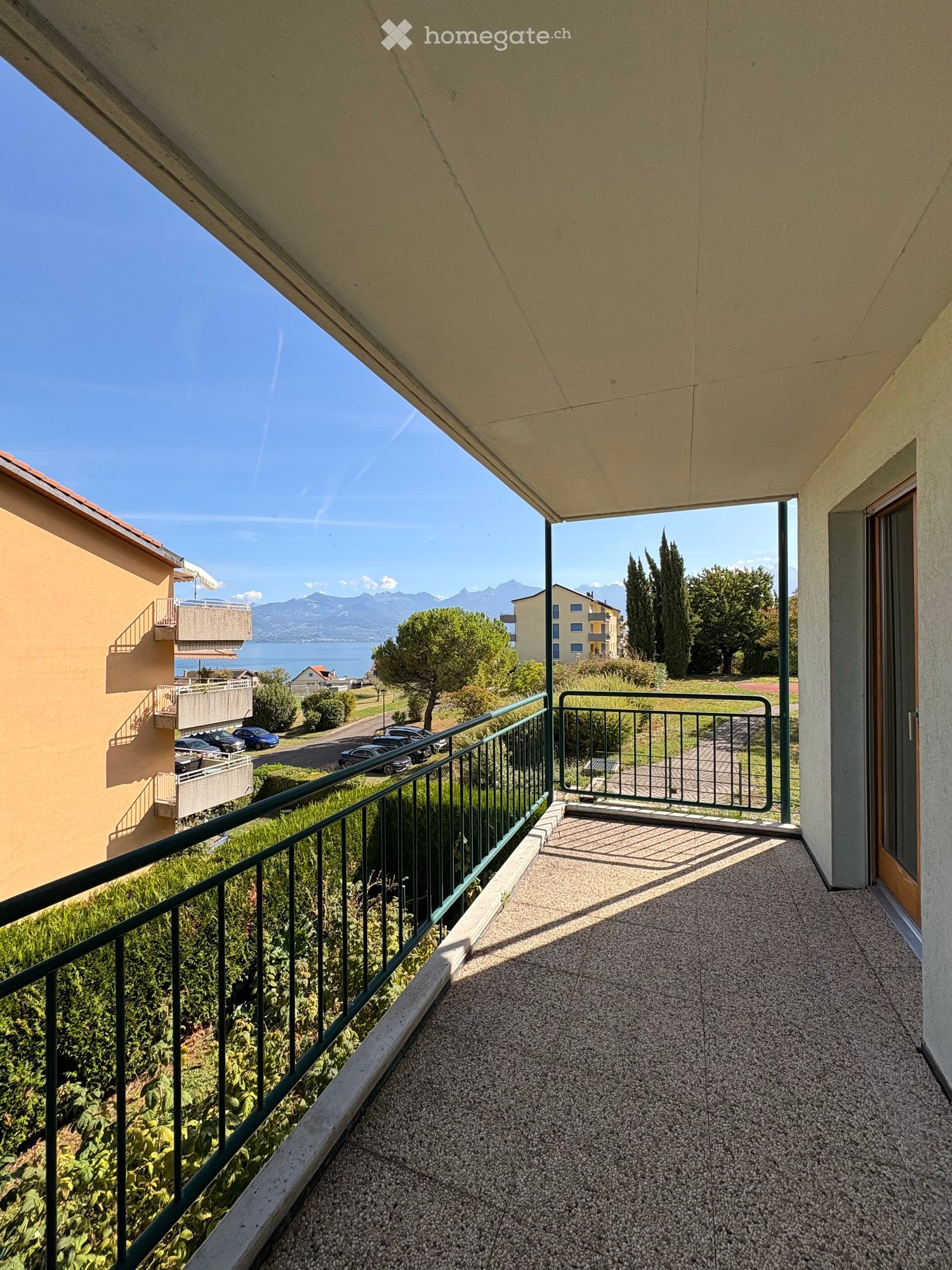 Balcony with metal railing, overlooking a scenic view of a lake and mountains, with surrounding greenery and parked cars visible.