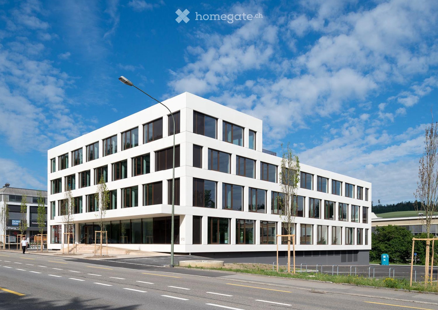 Modern, white, multi-story building with large windows and a clean, contemporary design. The building is situated on a street with a sidewalk and streetlight nearby.
