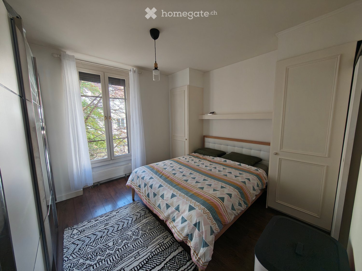 Cozy bedroom with a wooden bed, colorful bedspread, nightstand, wardrobe, and a large window with white curtains.