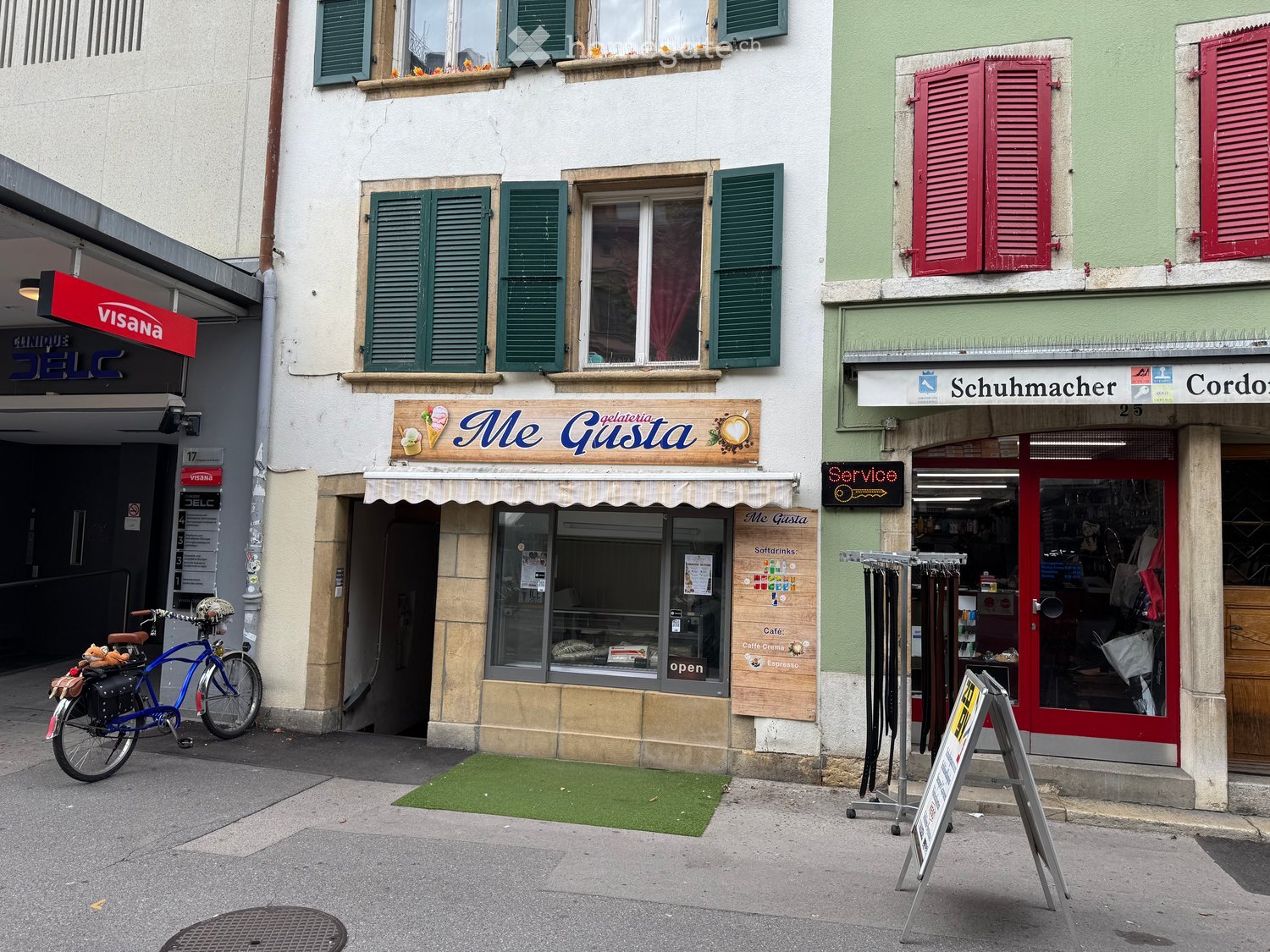 A street view showing a multi-story building with shops on the ground floor, including "Me Gusta" gelateria and a shoe shop. The building has green shutters and a mix of modern and traditional architectural elements.