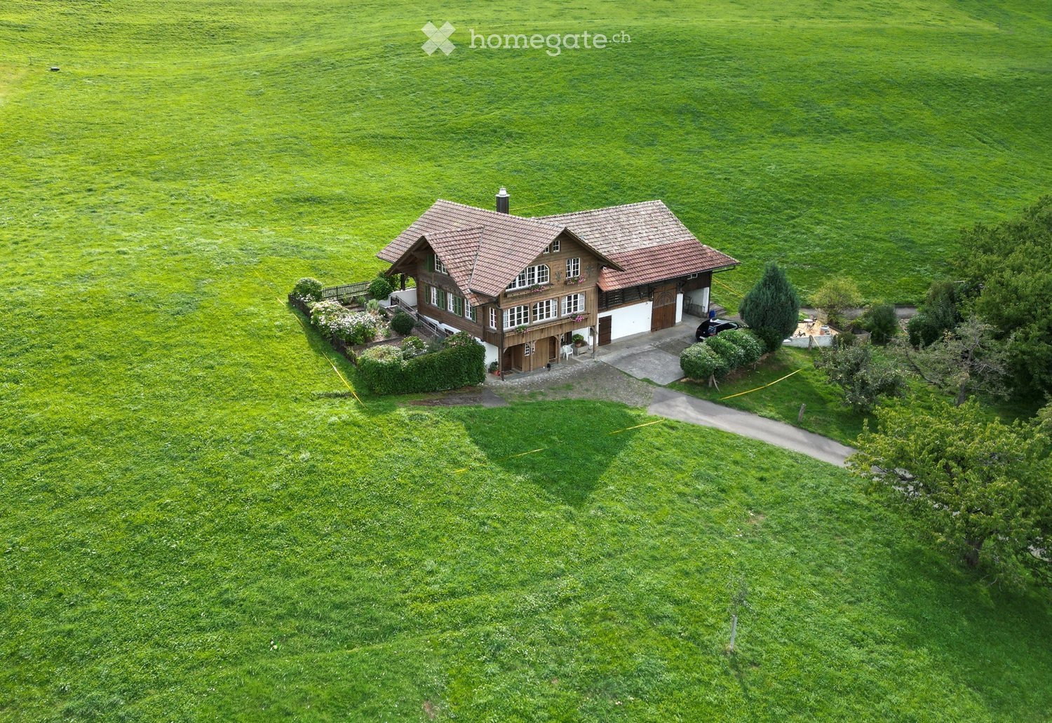 A two-story wooden house with a red-tiled roof, surrounded by a well-maintained lawn and garden, with a driveway and a parked car.