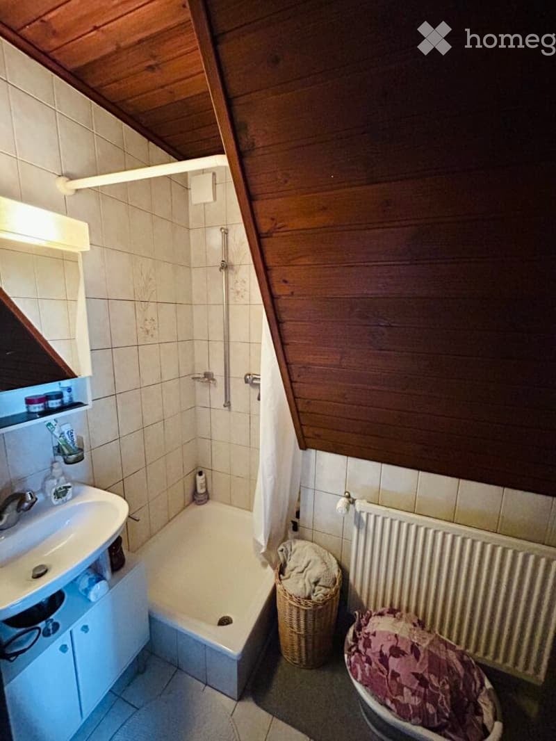 Bathroom with a wooden slanted ceiling, tiled walls, a white sink with a mirror, a bathtub, a radiator, and a wicker basket on the floor.