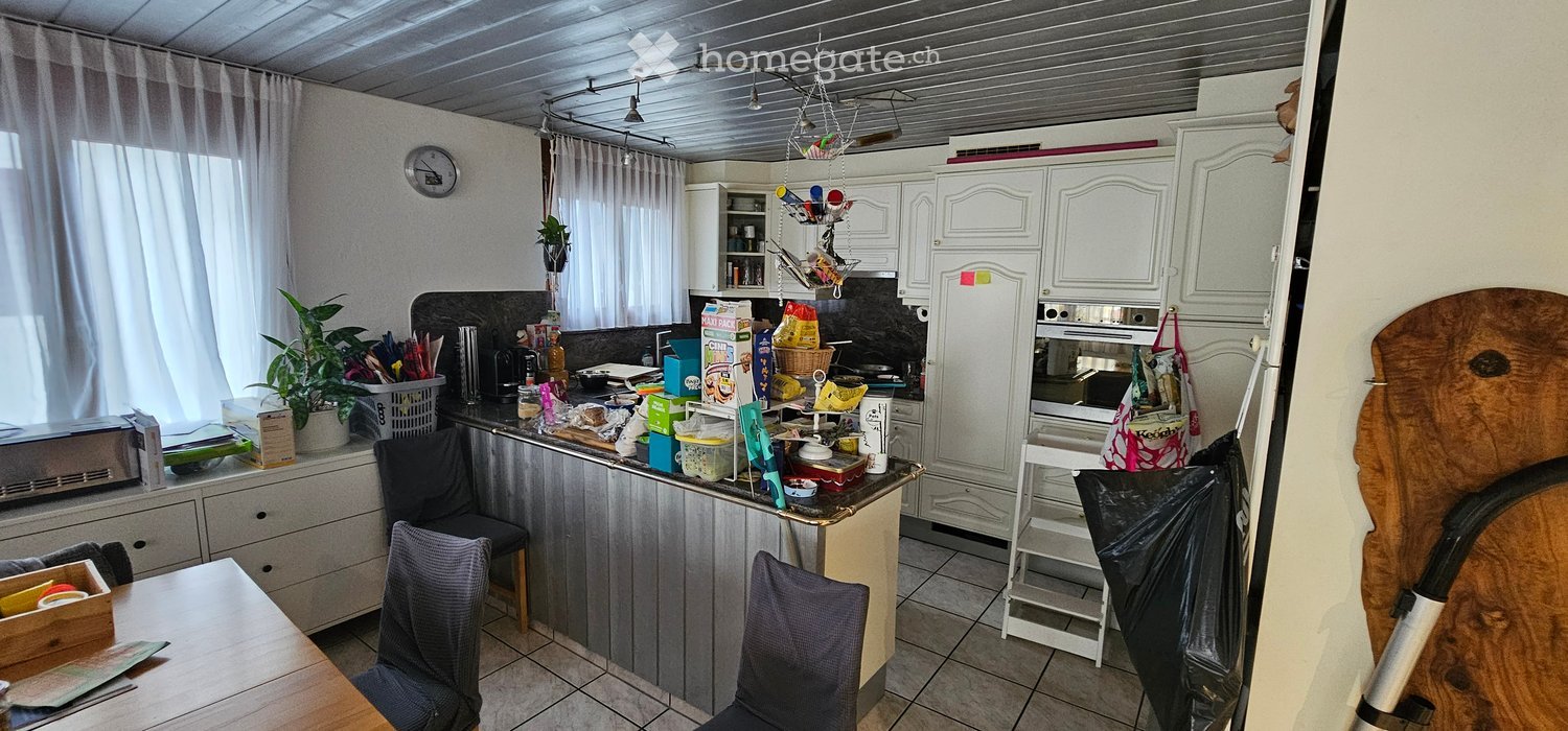 The kitchen features white cabinets, a refrigerator, an oven, and a microwave. There are chairs, a table, and a clock on the wall. The counter is cluttered with various items, including a basket, a plant, and some food packages.