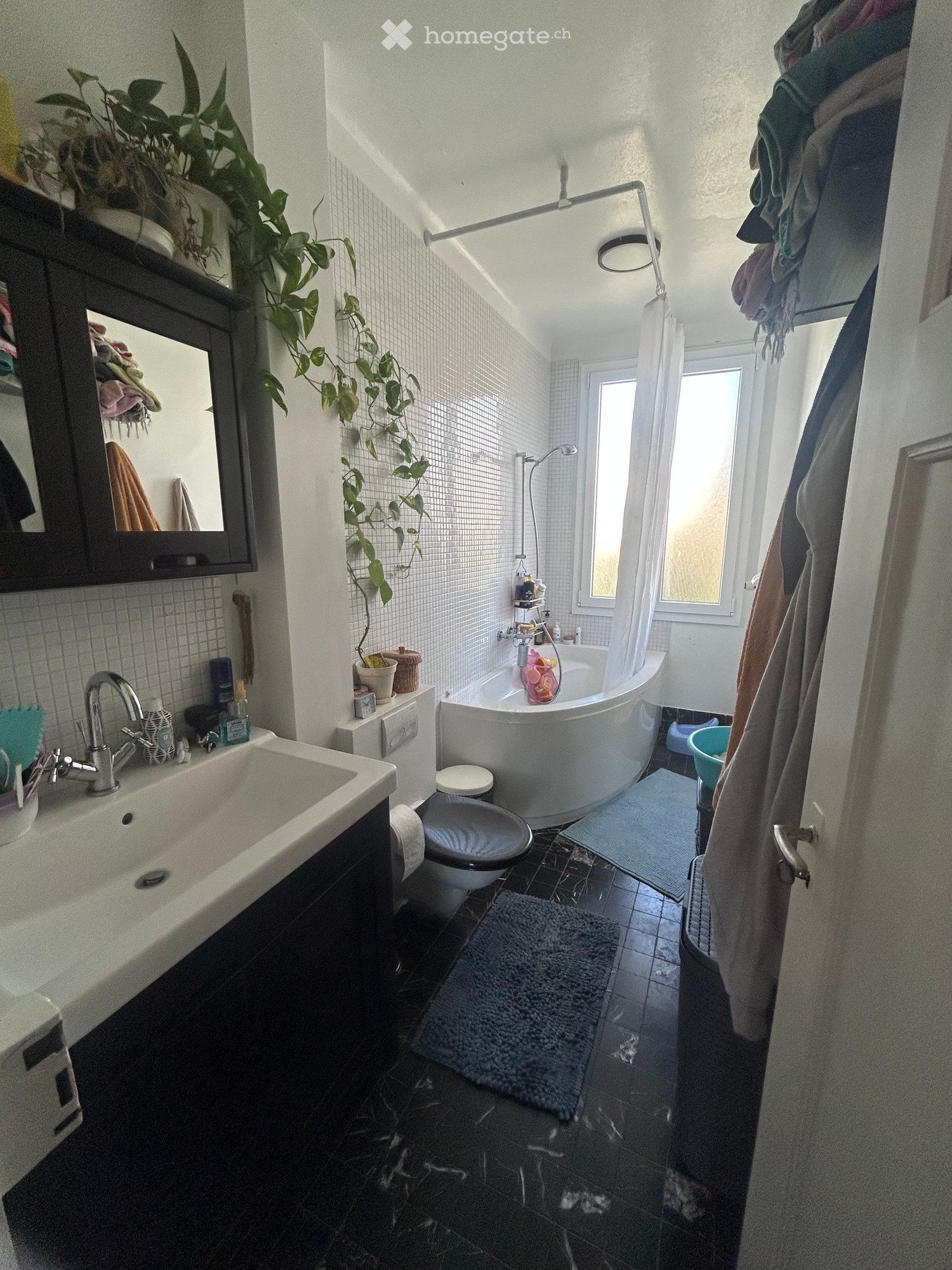 Modern bathroom with a white bathtub, toilet, sink, and mirror. Features a large window, a hanging plant, and a dark tiled floor.