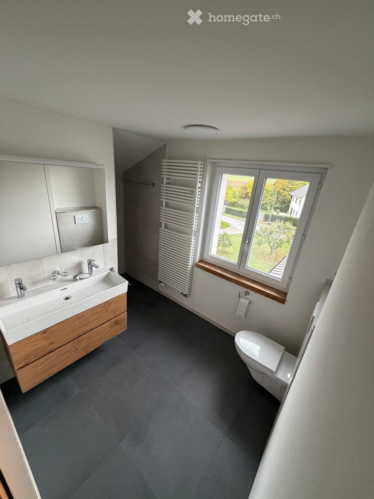 Modern bathroom with a double sink, wooden vanity, large mirror, radiator, and toilet. The room features dark grey tiles and a window with a view of the garden.