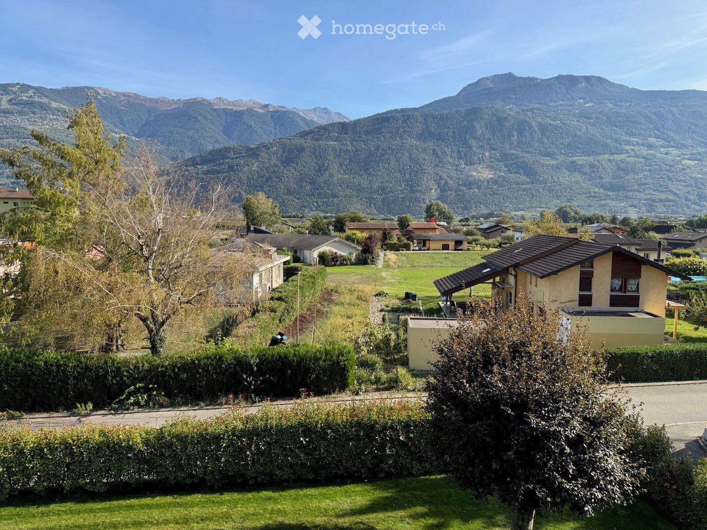 A peaceful residential area with houses, green fields, and a scenic mountain backdrop. The foreground features a well-maintained lawn and hedges, while the background showcases a picturesque valley with scattered houses and lush greenery.