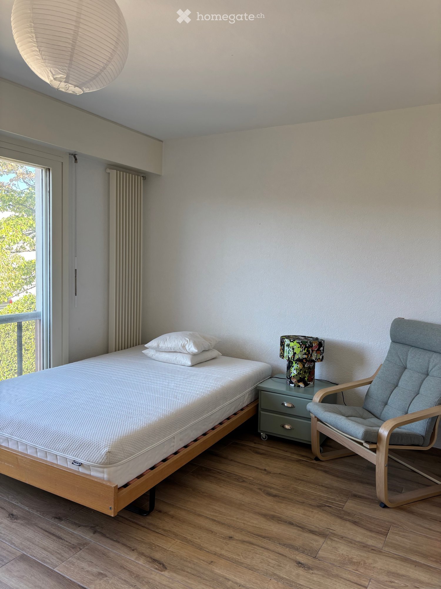 Modern bedroom with a wooden bed frame, white mattress, two pillows, a nightstand with a colorful lamp, and a wooden armchair. The room features light-colored walls, wooden flooring, and a large window with a view of greenery.