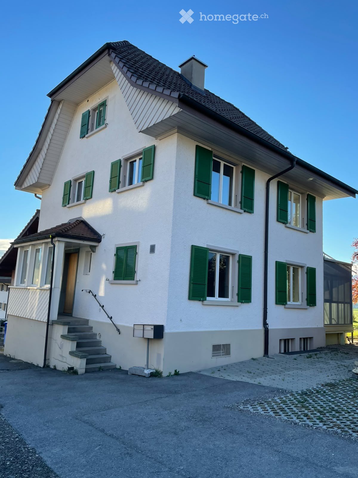 Two-story house with white walls, green shutters, and a brown tiled roof. Features include a front staircase, mailbox, and a small porch area.