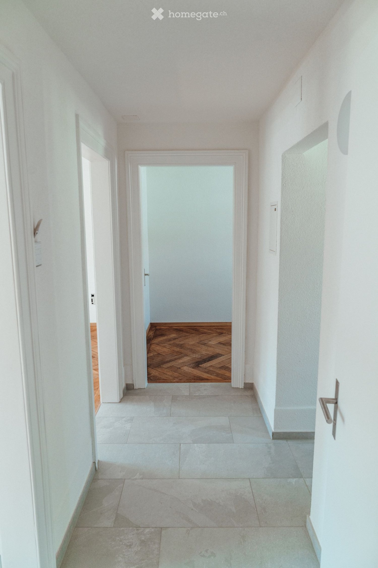 A clean, modern hallway with white walls, tiled flooring, and a wooden door at the end.
