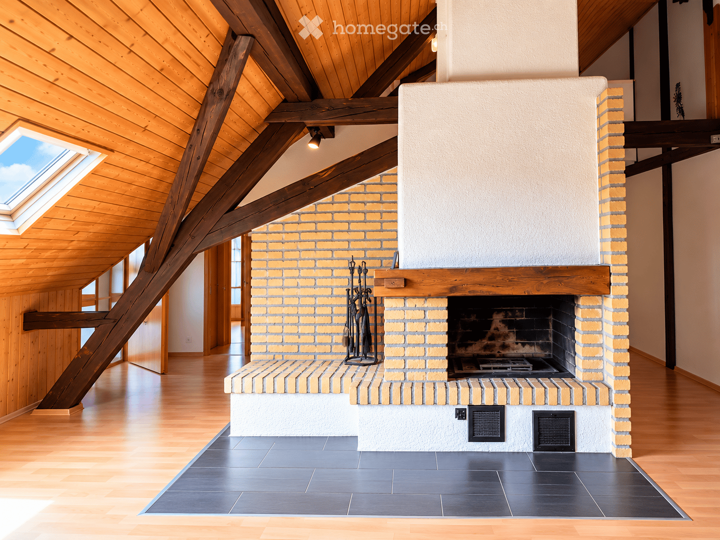 Modern living room with exposed wooden beams, skylight, and a brick fireplace with tools nearby.