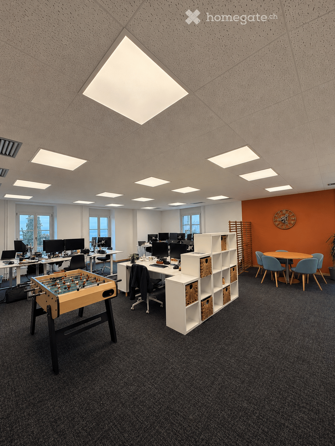 Modern office space with multiple workstations, a foosball table, and a round meeting table with chairs.