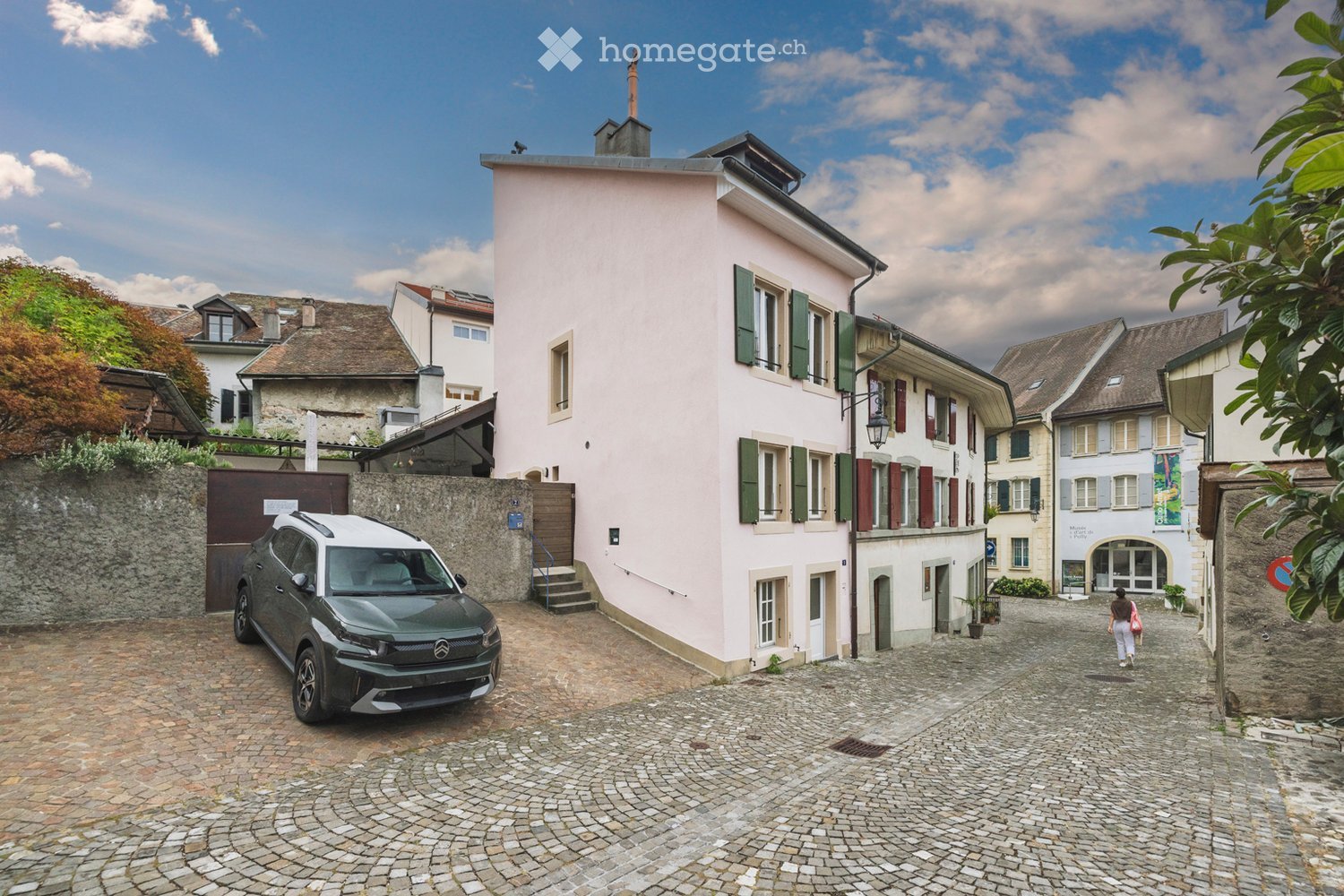 A charming pink building with green shutters and a stone staircase leading to the entrance, situated in a narrow cobblestone street with a parked car and surrounding historic architecture.