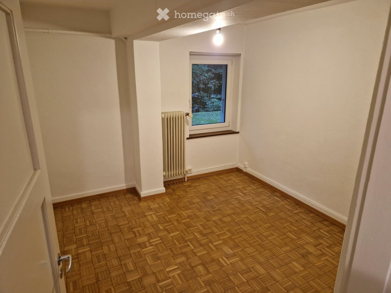 Empty room with wooden parquet flooring, white walls, a window with a view, and a radiator. No furniture or equipment present.