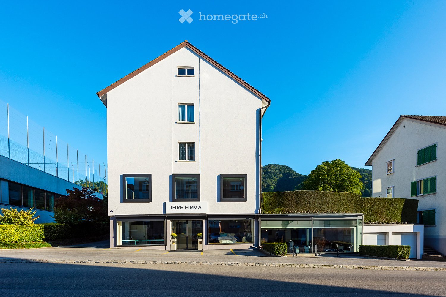 white facade, 3 windows, brown roof, name board 'IHRE FIRMA', front yard with bushes, glass entrance
