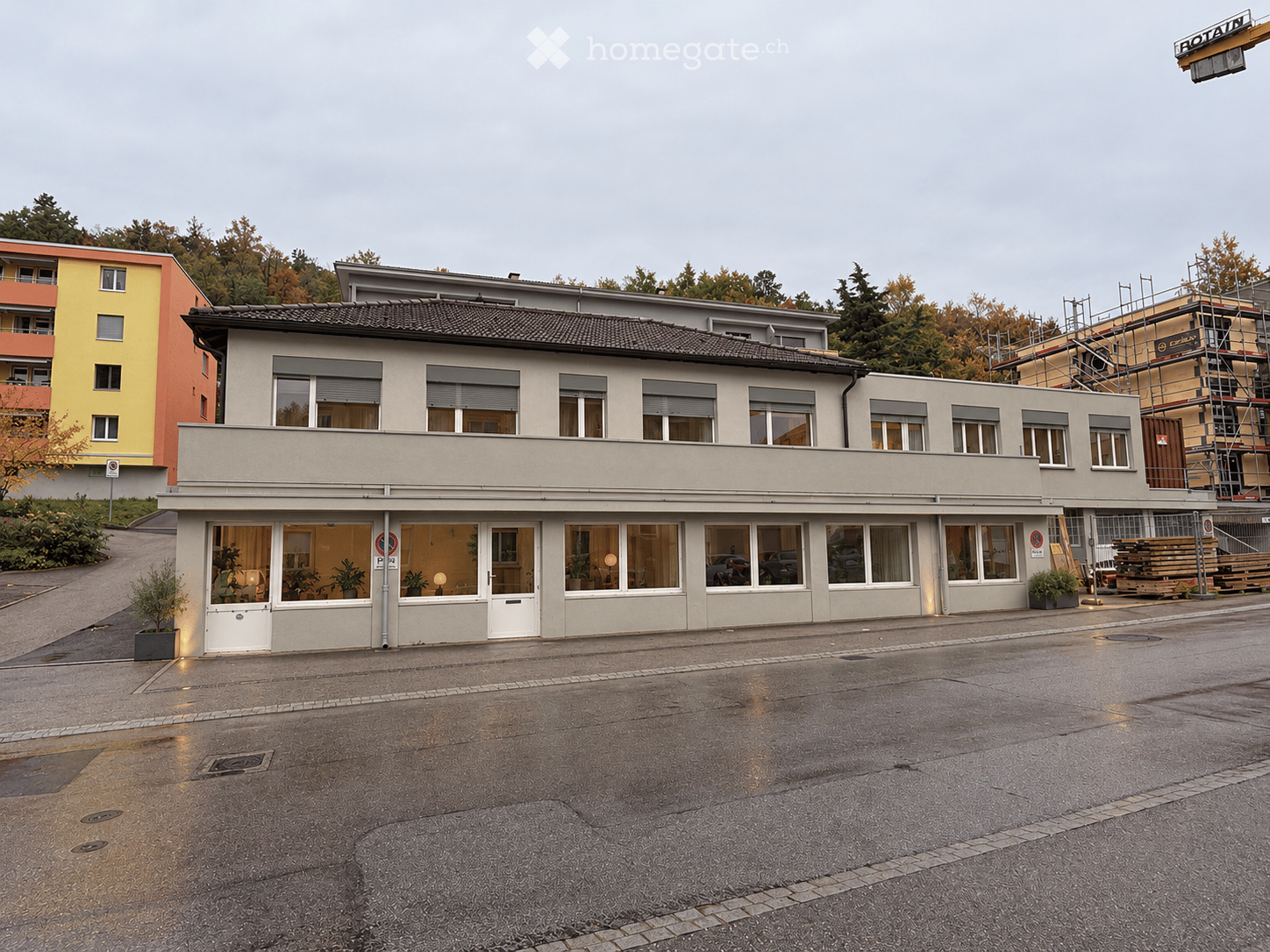 Two-story building with a flat roof and large windows, surrounded by trees and other structures.