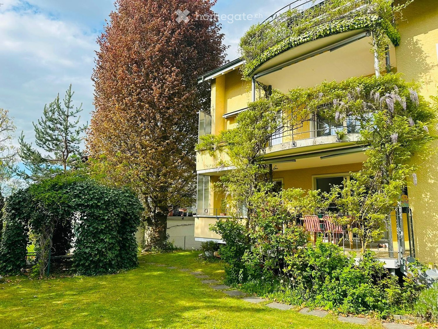 A well-maintained garden with a lawn, trees, and a multi-story yellow building with balconies and greenery on the facade.