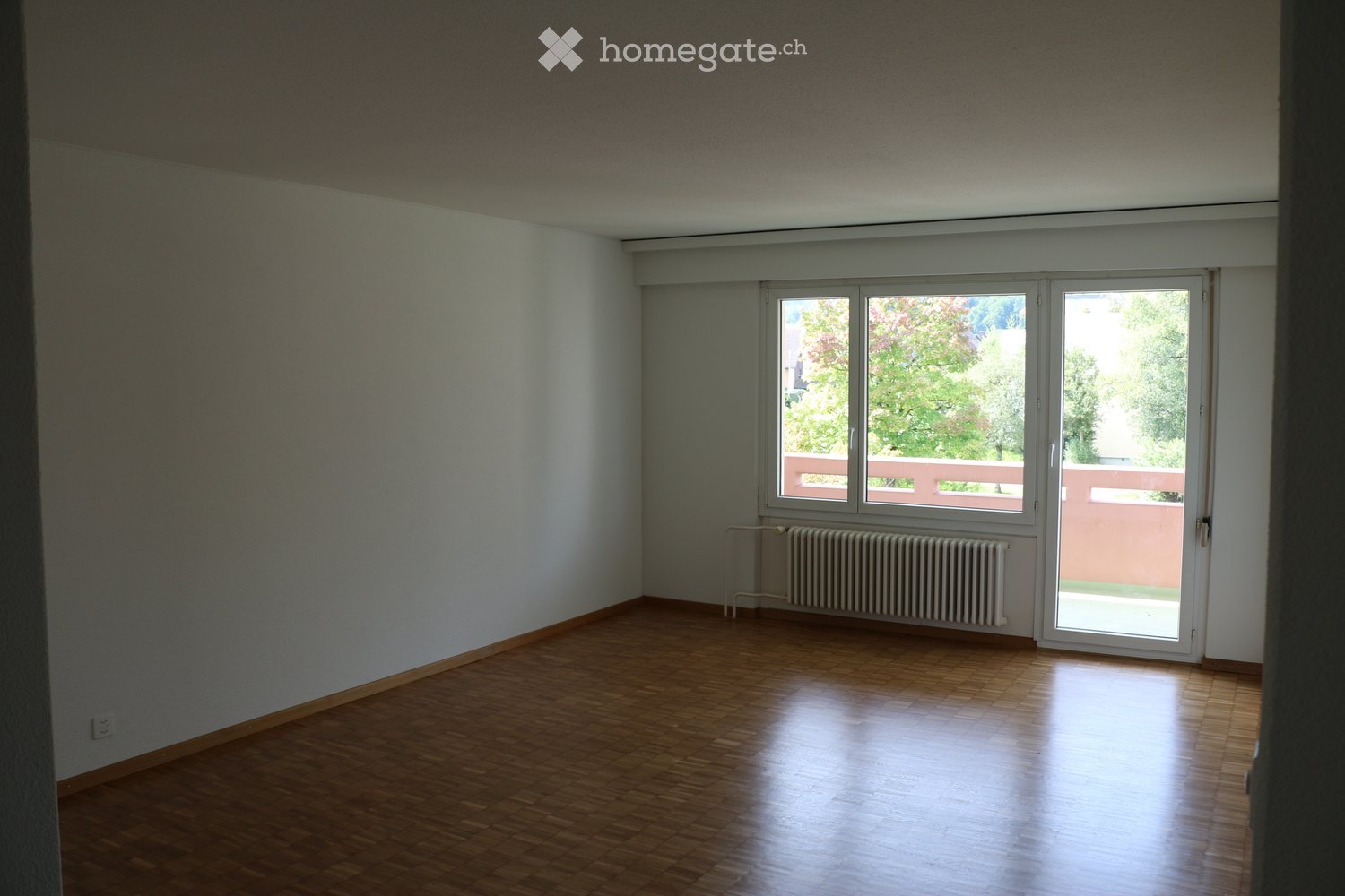 Empty room with wooden flooring, white walls, large windows, and a radiator. No furniture or equipment present.