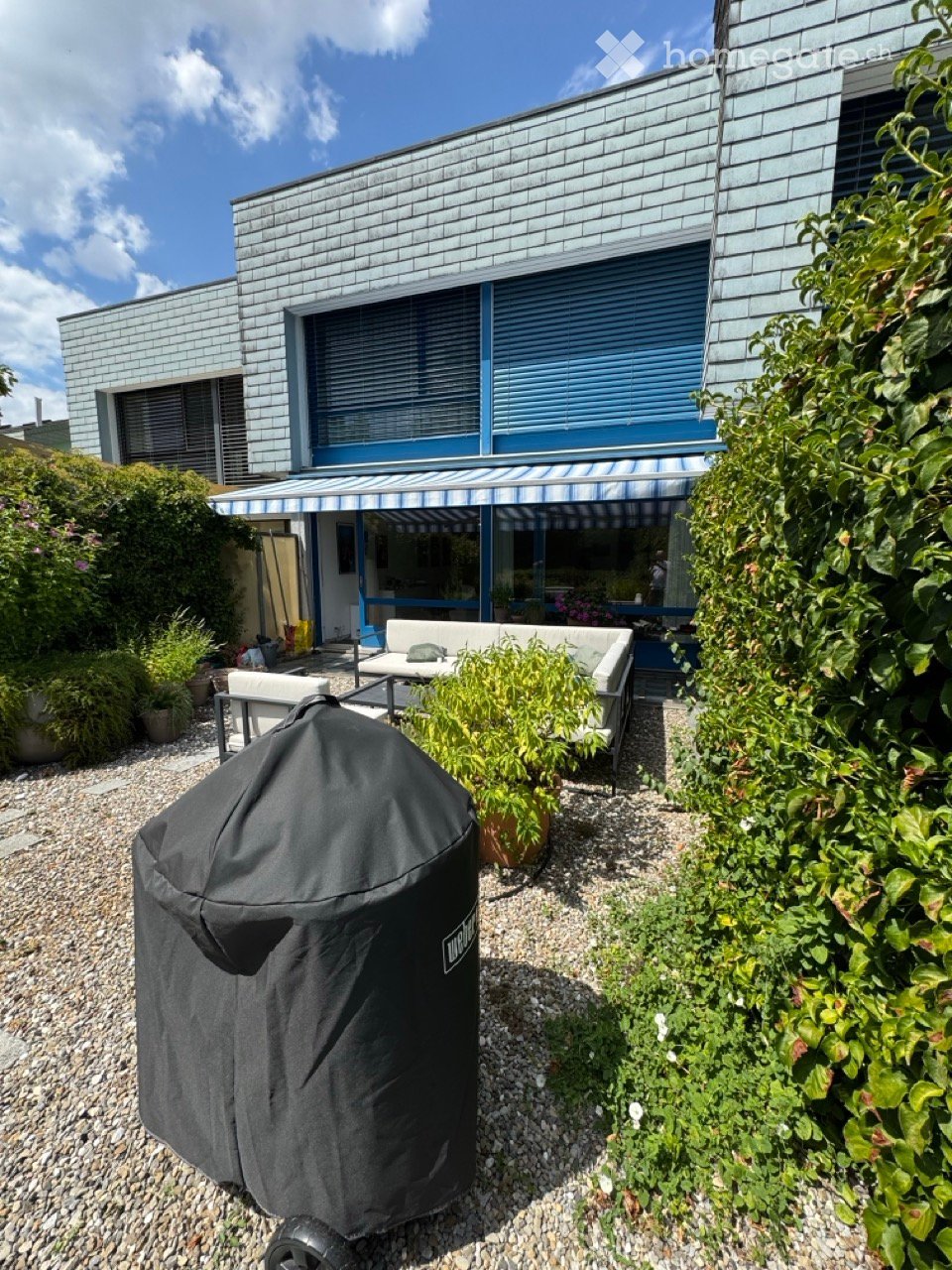 Outdoor patio area with a covered seating area, featuring a sofa, table, chairs, and a covered grill, surrounded by plants and a modern house with blue shutters.