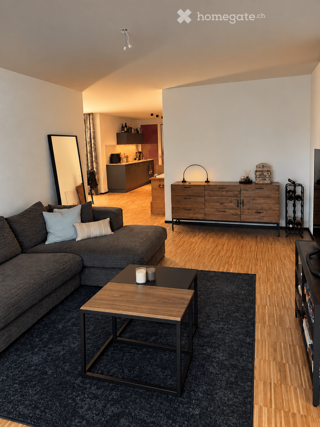 Modern living room with a sectional sofa, coffee table, large mirror, wooden dresser, wine rack, and a visible kitchen area with cabinets and appliances.