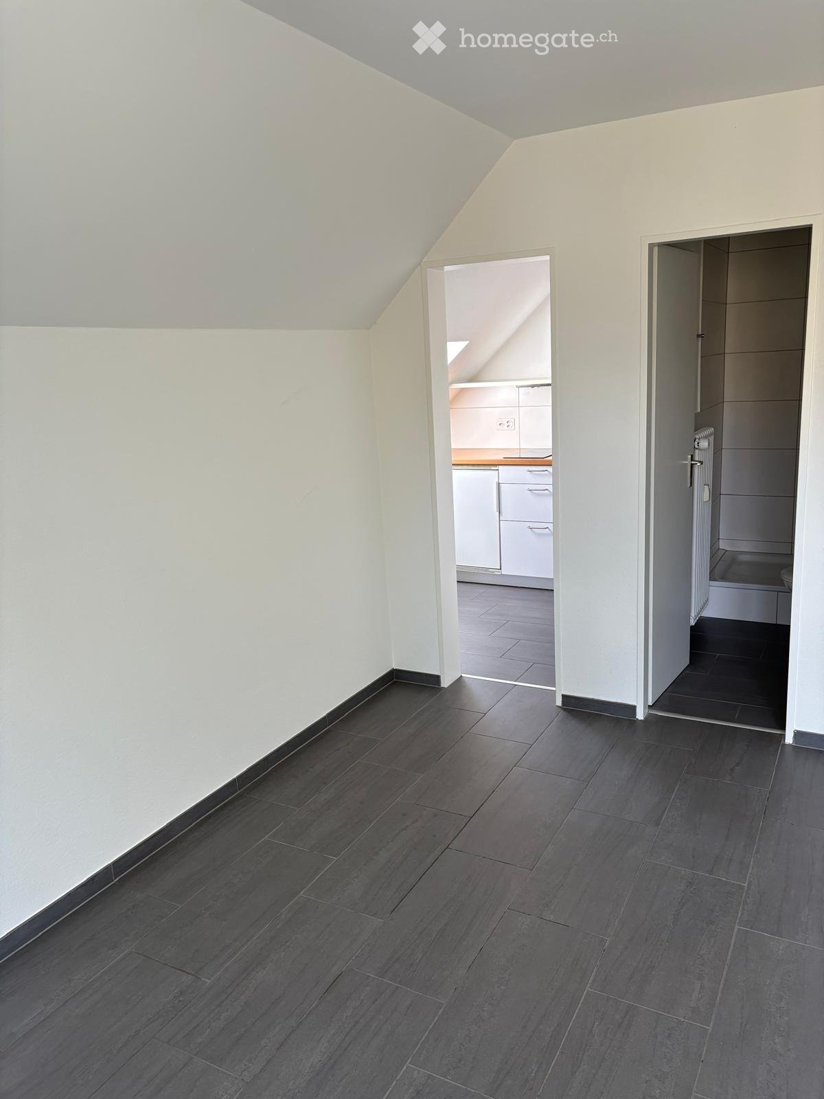 Empty room with dark grey tiled flooring and white walls, featuring an open doorway to a kitchen and another doorway to a bathroom.