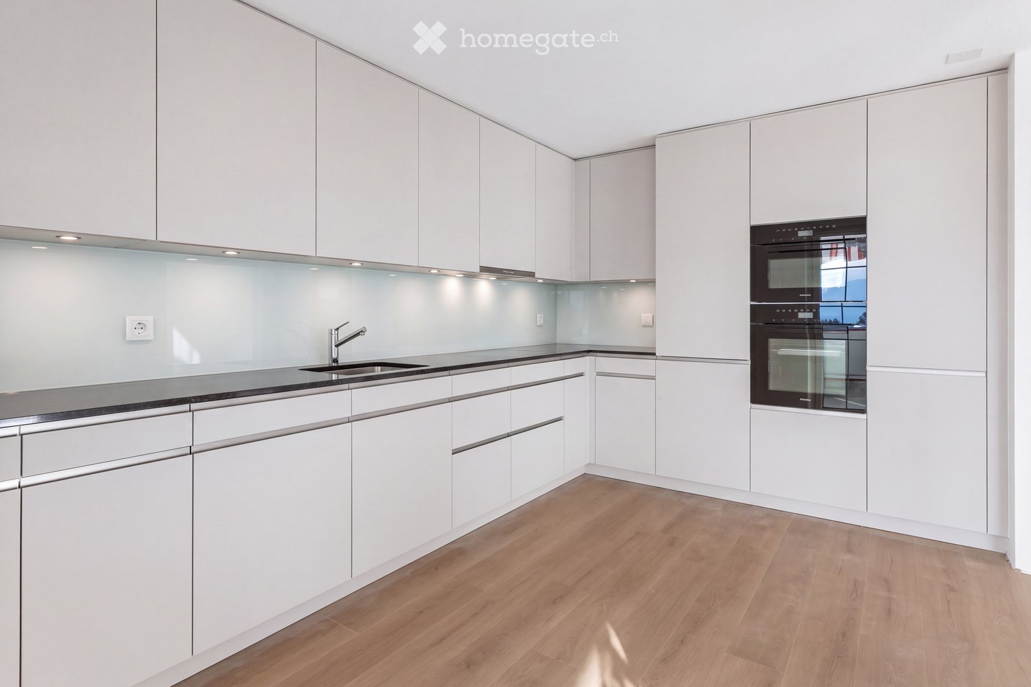 Modern kitchen with white cabinets, wooden flooring, stainless steel sink, and built-in oven.