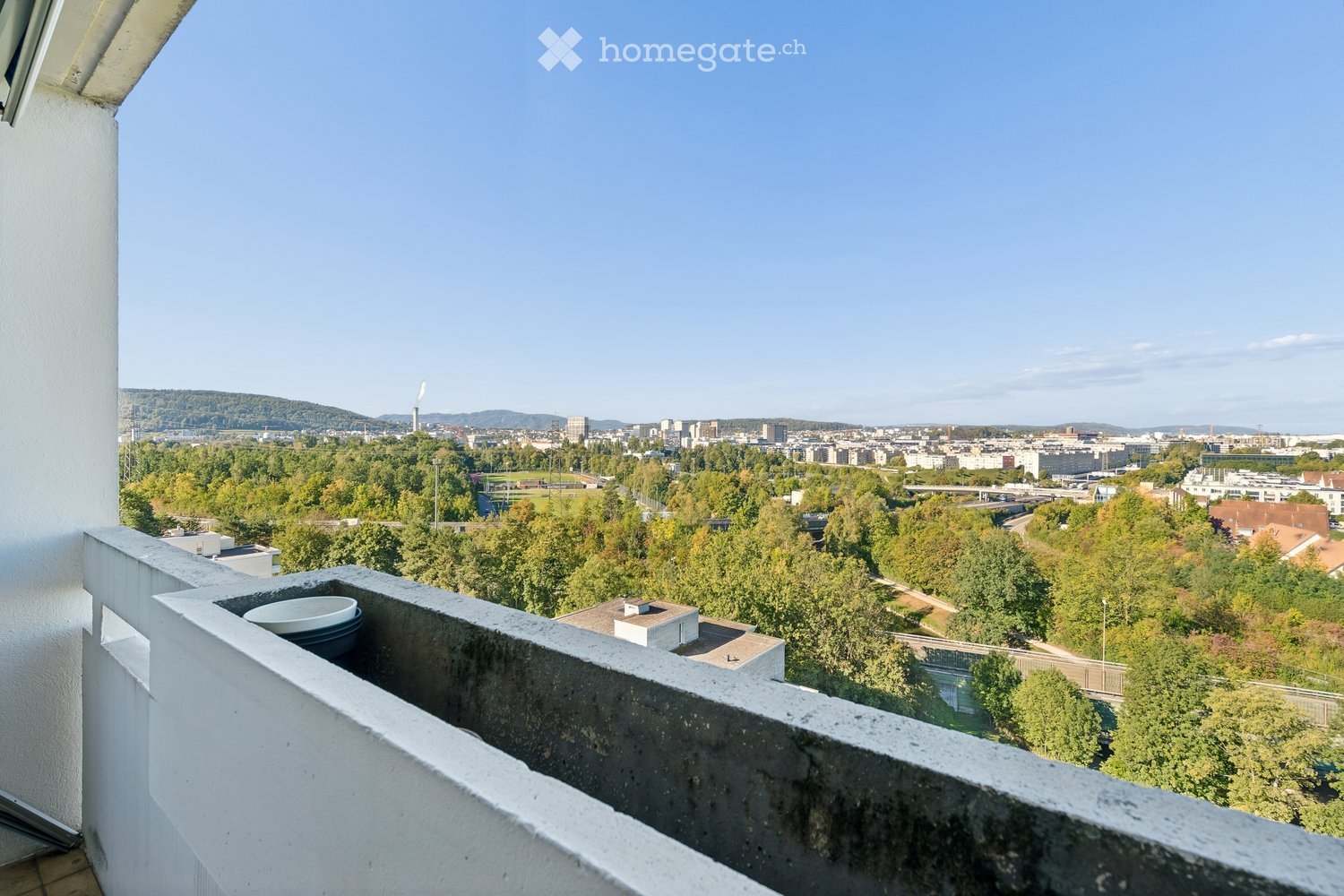 Balcony with a concrete railing, overlooking a cityscape with trees, buildings, and distant hills under a clear blue sky.