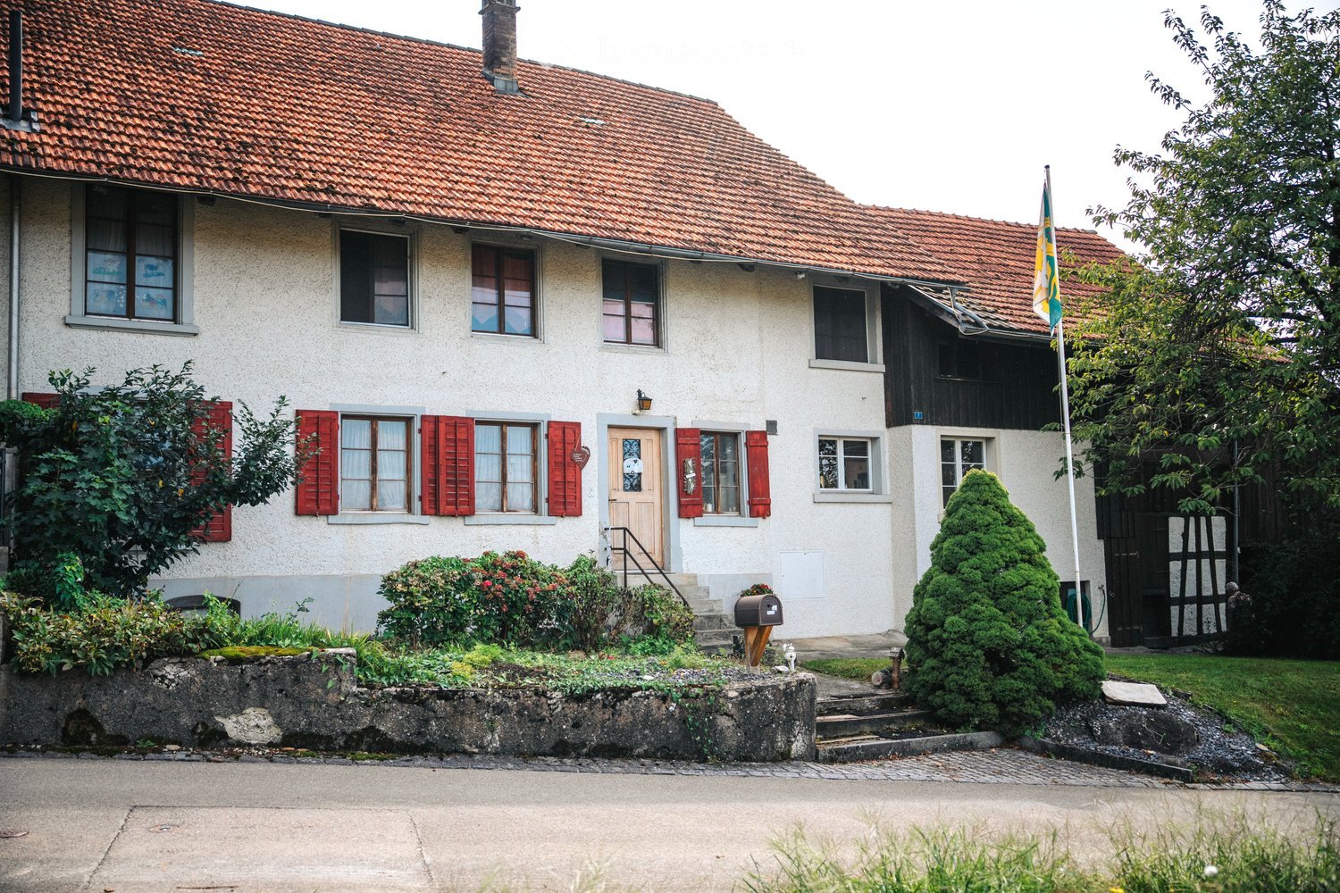 Two-story house with red-tiled roof, white walls, red shutters, and a small garden with shrubs and a flagpole.