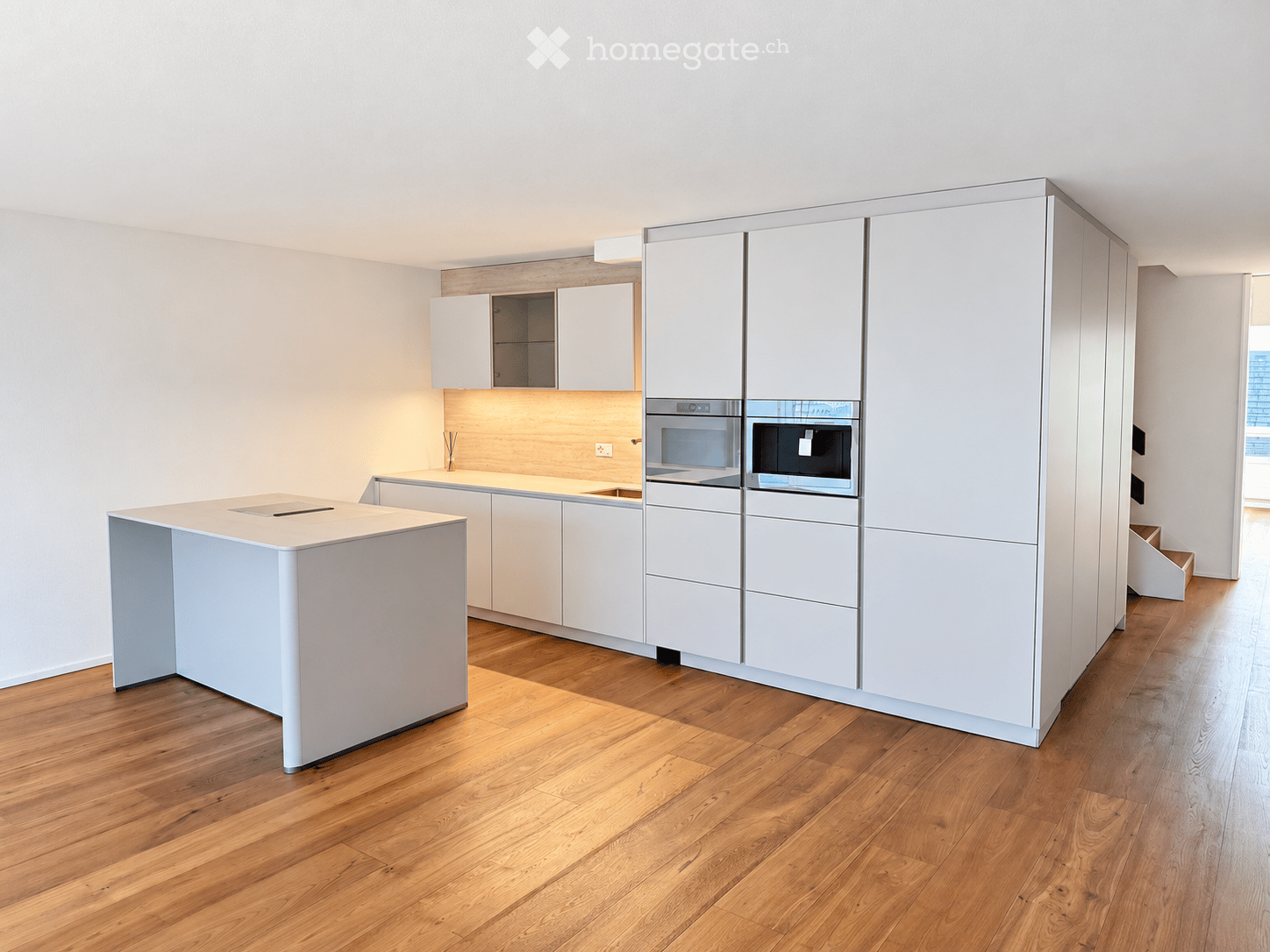Modern kitchen with white cabinets, a central island, and integrated appliances including an oven and microwave. The floor is made of light wood, and the walls are painted white.