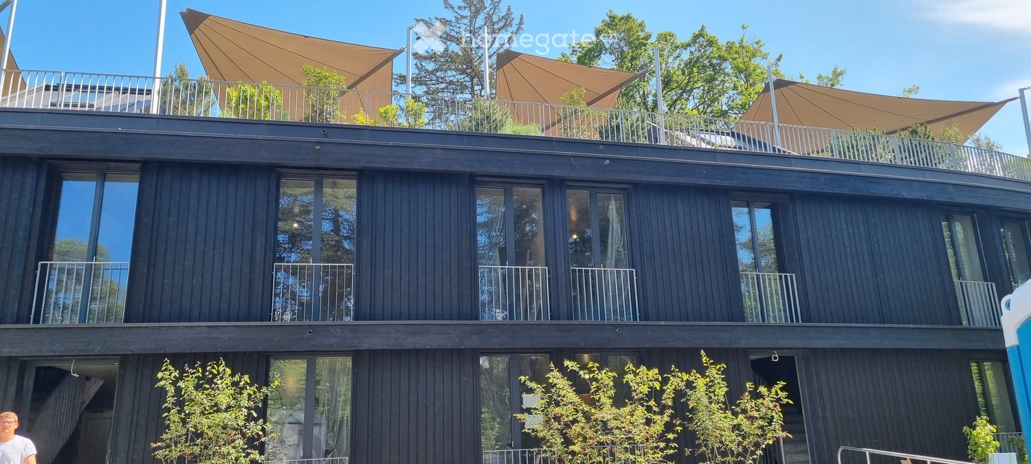 Modern two-story building with dark wooden facade, large windows, and multiple balconies. The roof features a large canopy and additional seating area.