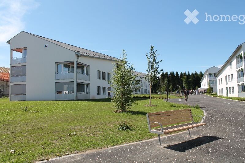Modern white multi-story building with large windows and balconies, surrounded by green lawn and trees. A wooden bench is placed on the pathway in front of the building.