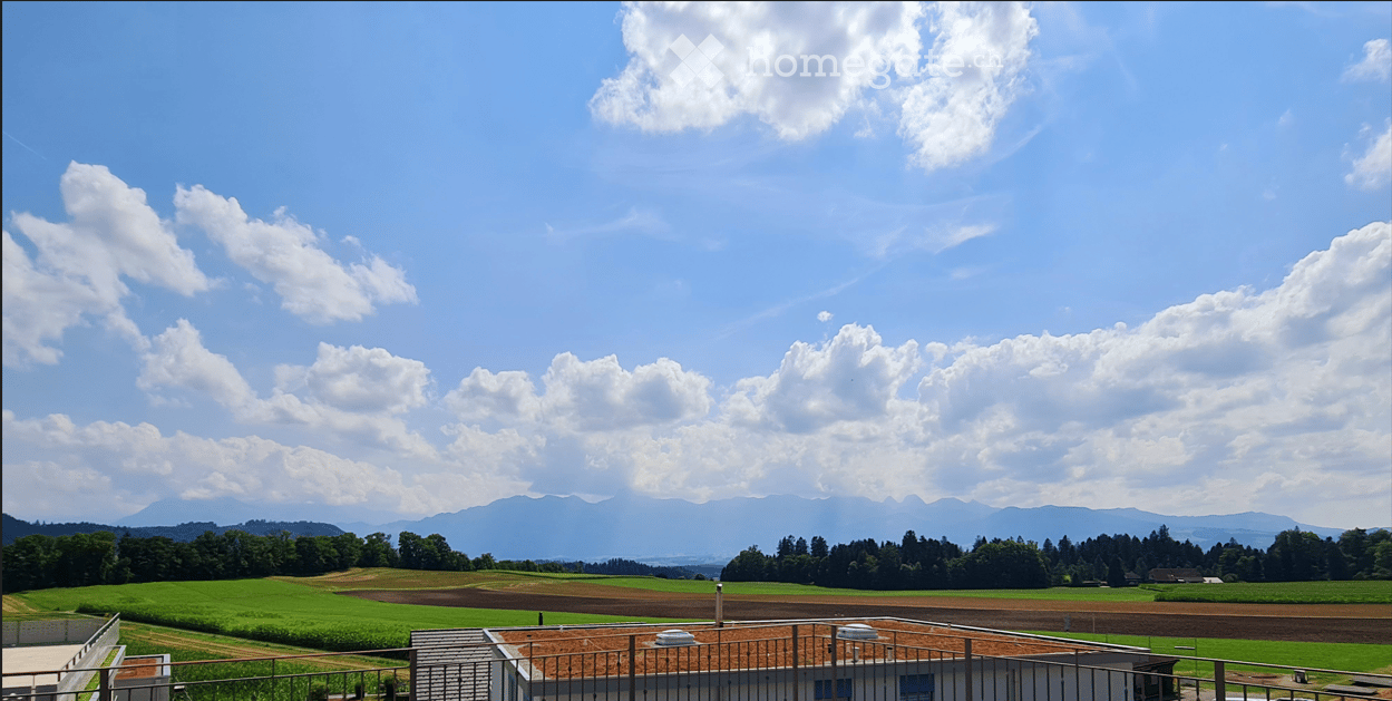 A scenic view of a rural landscape with green fields, scattered trees, and distant mountains under a partly cloudy sky.