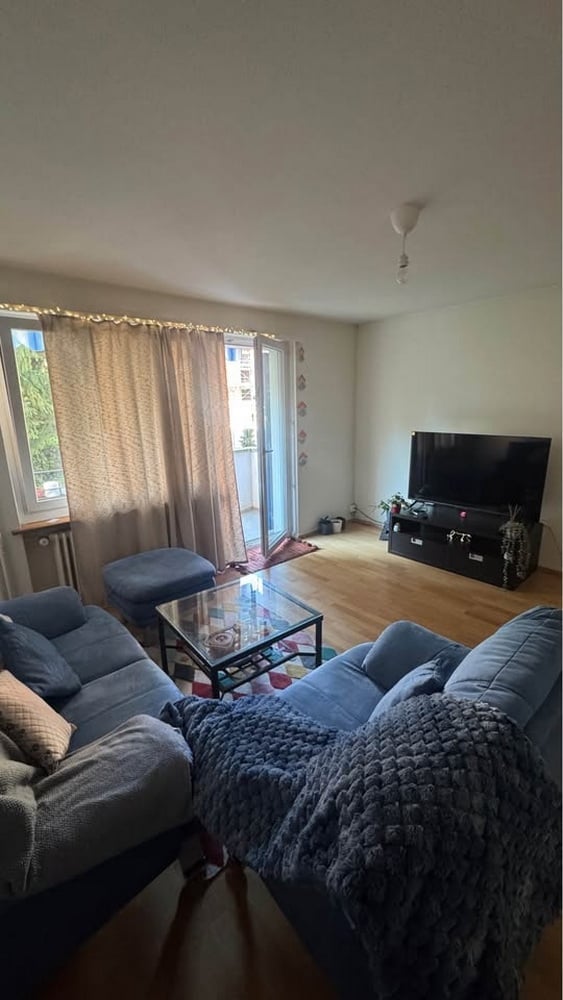 Modern living room with wooden flooring, large windows, and a balcony access. Features a grey sectional sofa, glass coffee table, ottoman, TV stand, and floor lamp.