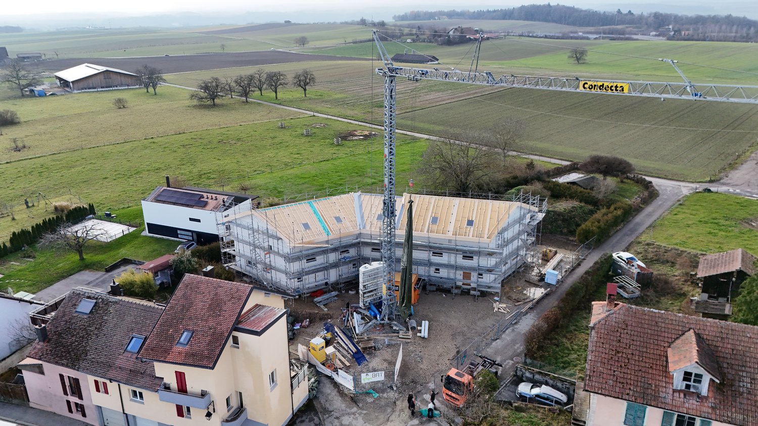An ongoing house construction site with a large crane, surrounded by green fields and a few neighboring houses, one of which has solar panels on the roof