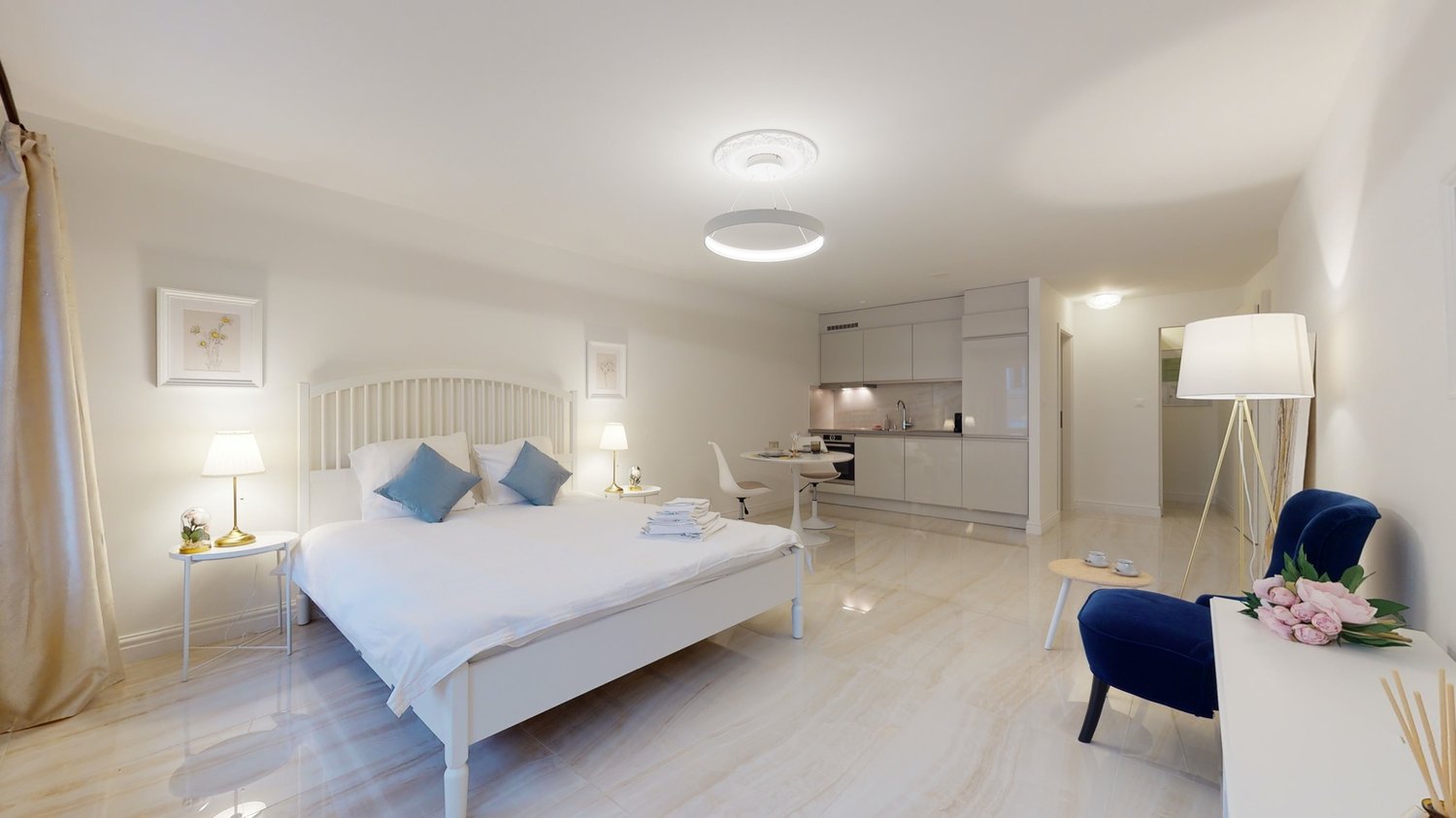 Bed with white sheets and blue pillows, bedside tables with lamps, a blue chair, a kitchen with white cupboards, open floor design, two lamps and a table