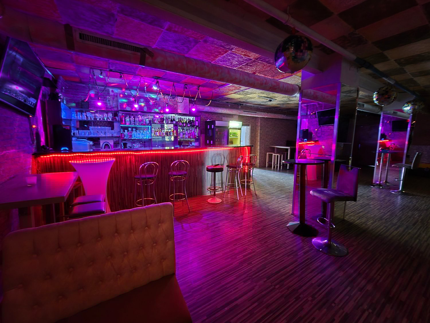 Dimly lit bar with purple and red lighting, a long bar with stools, couches and tables, a disco ball hanging from the ceiling, and various liquor bottles and glassware on the shelves behind the bar