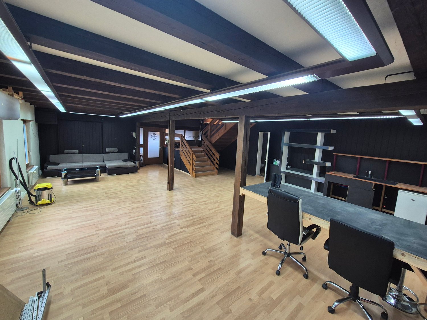 Large open office space, hardwood floors, wooden beams, office chairs, tables, couch, wooden staircase, ventilation units
