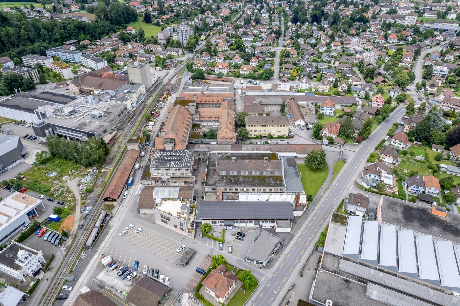 urban landscape with houses, roads, and buildings. A central area with several buildings, surrounded by other structures, including residential homes, commercial properties, and industrial facilities. Green areas, including lawns and trees, are scattered