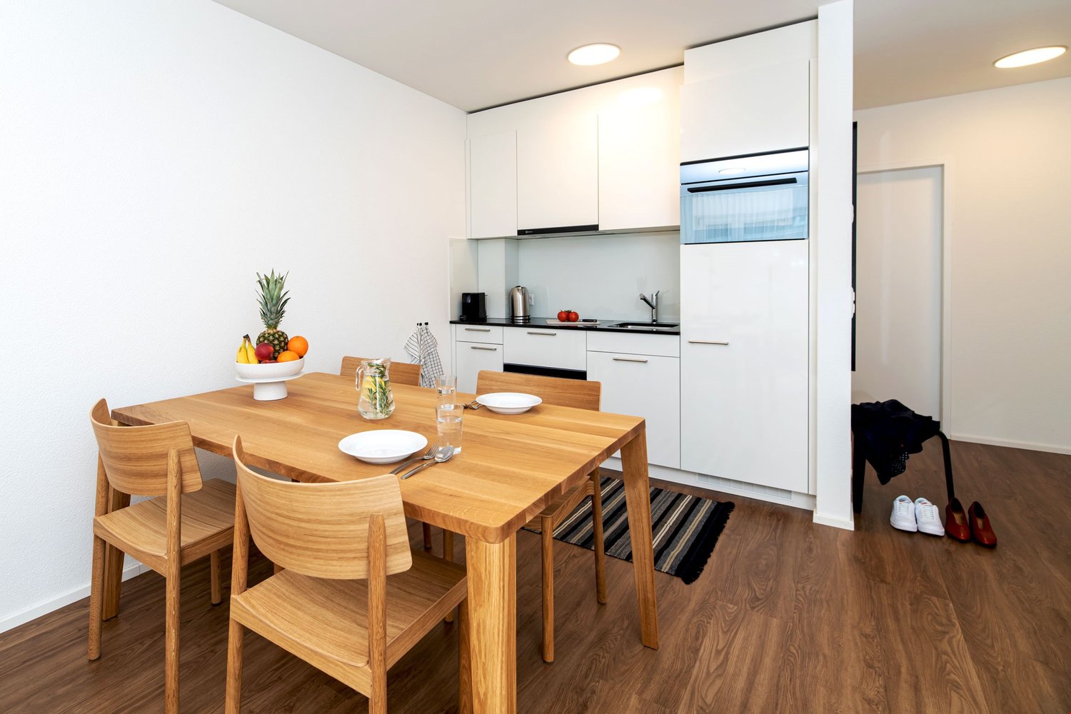 A wooden dining table with 4 chairs, white kitchen with oven, fridge, and sink