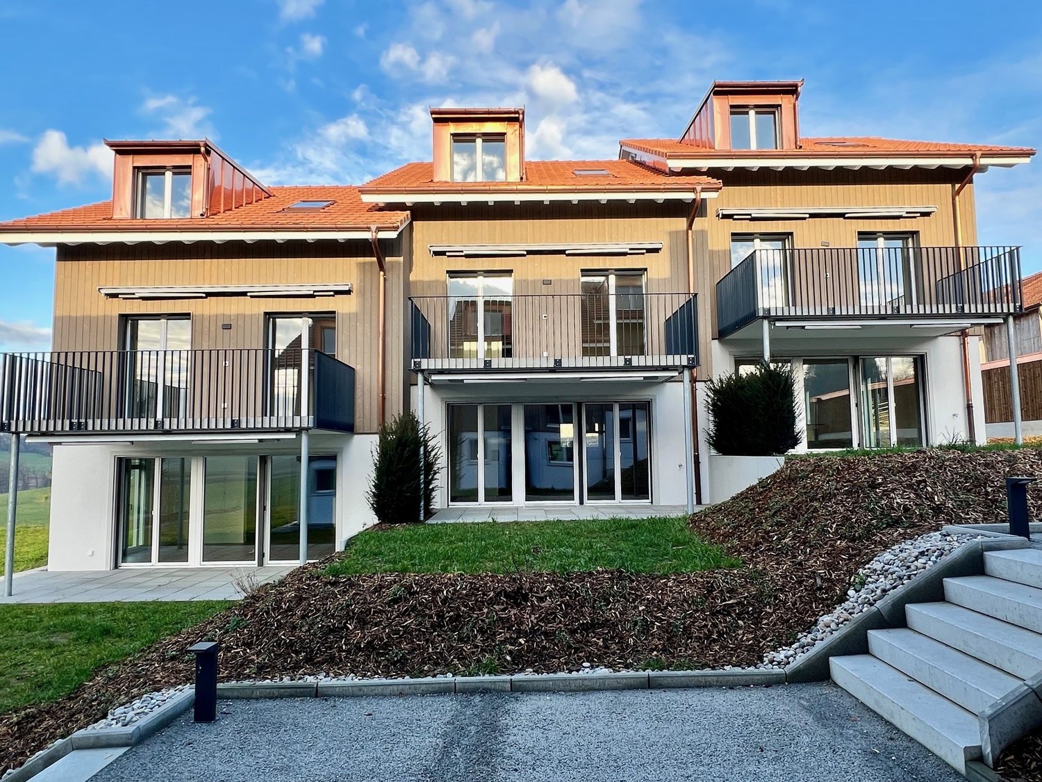 Two-story building, modern architecture, red roof, glass windows, large balconies on both floors, well-maintained garden in front, large steps leading to the entrance, concrete driveway.